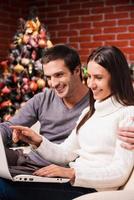 Buying online. Beautiful young couple using computer together while woman pointing monitor and smiling with Christmas Tree in the background photo