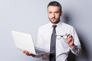 Confident business expert. Confident mature man in shirt and tie holding laptop and eyewear while standing against grey background photo