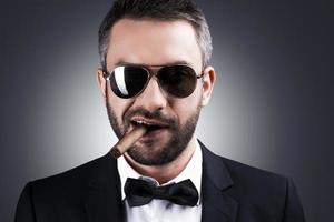 Just me and my cigar. Portrait of handsome mature man in formalwear and sunglasses smoking cigar and smiling while standing against grey background photo