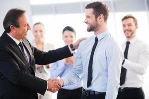 Good job Two cheerful business men shaking hands while their colleagues applauding and smiling in the background photo