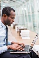 Businessman at work. Side view of young and confident African man in formalwear working on laptop while sitting at his working place photo