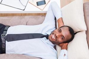 Relaxing after long day working. Top view of handsome young African man in shirt and tie holding hands behind head and smiling while lying on the couch photo