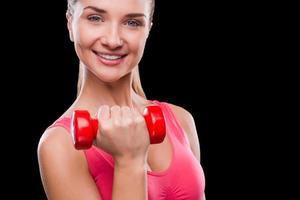 Keep your body fit Sporty young woman exercising with dumbbell and smiling while standing against black background photo