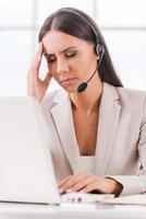 Sick and tired. Depressed young businesswoman in headset touching her head with hand and keeping eyes closed while sitting at her working place photo
