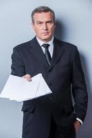 I got some paperwork for you. Confident mature man in formalwear stretching out documents and looking at camera while standing against grey background photo