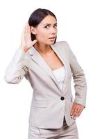 I love gossip Surprised young businesswoman in suit holding hand near ear while standing against white background photo