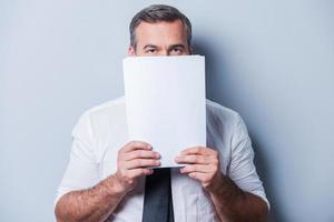 Stuck in paperwork. Confident mature man in formalwear hiding part of his face behind documents and looking at camera while standing against grey background photo