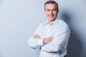 Confident mature man. Portrait of confident mature man in shirt looking at camera and smiling while keeping arms crossed and standing against grey background photo