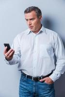 Shocking news. Surprised mature man in shirt holding mobile phone and looking at it while standing against grey background photo