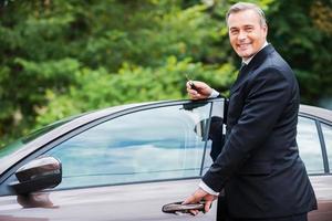 este coche es perfecto para mí. hombre maduro alegre en ropa formal mirando a la cámara y sonriendo mientras está de pie cerca de su coche nuevo foto