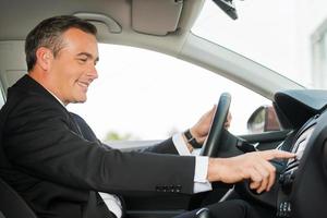 Driving with comfort. Side view of cheerful mature man in formalwear driving car and touching dashboard with finger photo