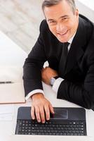 Businessman at work. Top view of happy mature man in formalwear working on laptop and smiling while sitting at his working place photo