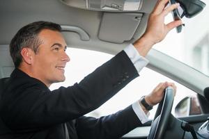 Examining his new car. Side view of cheerful mature man in formalwear adjusting mirror while sitting in his car photo