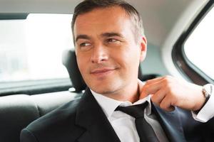 Relaxing after business trip. Satisfied mature businessman adjusting his necktie while sitting on the back seat of a car photo