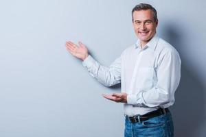 Selling your product. Confident mature man in shirt looking at camera and pointing away while standing against grey background photo