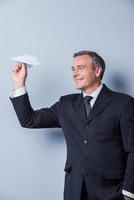 Playful businessman. Cheerful mature man in formalwear holding paper airplane and smiling while standing against grey background photo