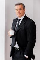 Taking time for coffee break. Confident mature man in formalwear holding coffee cup and looking at camera while standing near the window photo