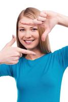 Focusing at you. Beautiful young woman looking at camera and gesturing finger frame while standing against white background photo