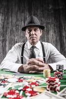 Serious senior man in shirt and suspenders sitting at the poker table and looking at camera with cards  with money and  gambling chips laying all around him photo