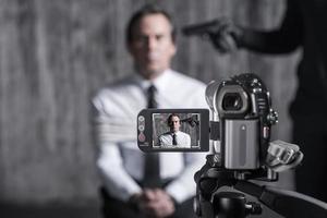 Filming hostage. Tied up businessman caught by a criminal sitting in front of a dirty wall with gun near his head while video camera filming it on the foreground photo