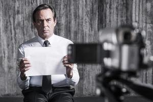 Hopeless hostage. Frustrated businessman caught by a criminal sitting in front of a dirty wall and holding paper while video camera filming it on the foreground photo