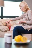 mamá cuidará de ti. madre sosteniendo un termómetro y tocando la frente de su hija enferma tendida en el sofá foto