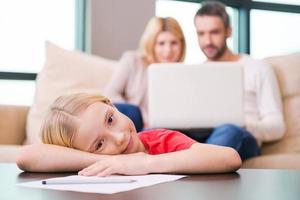 tiempo libre en familia. niña feliz apoyando su rostro en la mesa y sonriendo mientras sus padres se sientan en el fondo con una laptop foto