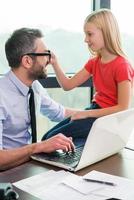 I need your glasses Cheerful father in shirt and tie working on laptop while his playful daughter sitting close to him and touching his eyeglasses photo