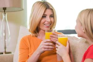 nos encanta el jugo de naranja. feliz madre e hija sosteniendo vasos con jugo de naranja y mirándose con una sonrisa mientras se sientan juntos en el sofá foto