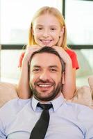 yo y mi padre. linda niñita uniéndose a su alegre padre sentado en el sofá de casa foto