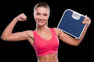 In perfect form. Sporty young woman holding weight scale and showing her bicep while standing against black background photo
