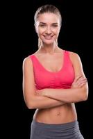 Sporty beauty. Sporty young woman keeping arms crossed and smiling while standing against black background photo