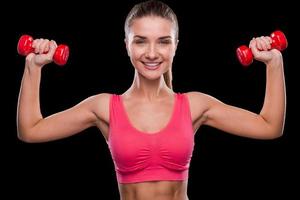 Beautiful and sporty. Sporty young woman exercising with dumbbell and smiling while standing against black background photo