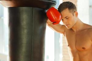 sentirse cansado después del entrenamiento. joven boxeador cansado con guantes deportivos apoyado en el saco de boxeo foto