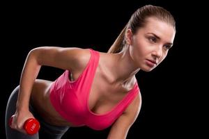 Training her body. Sporty young woman exercising with dumbbell while standing against black background photo