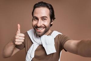 joven feliz haciendo selfie y mostrando su pulgar hacia arriba mientras está de pie contra el fondo marrón foto