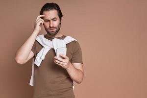 joven frustrado mirando un teléfono inteligente mientras se enfrenta a un fondo marrón foto