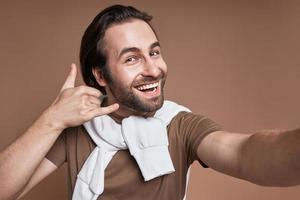 Handsome young man making selfie and gesturing while standing against brown background photo