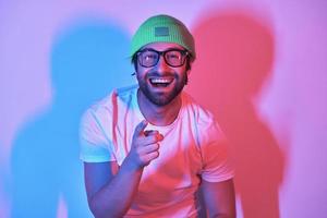 Un joven feliz con un sombrero funky apuntando a la cámara mientras se enfrenta a un fondo colorido foto