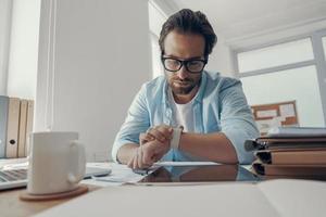 Handsome young man looking at his smart watch while sitting at his working place in office photo