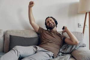 joven feliz con auriculares sosteniendo un teléfono inteligente y gesticulando mientras se relaja en el sofá foto