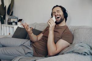Cheerful young man in headphones using smart phone like microphone while relaxing on the couch photo