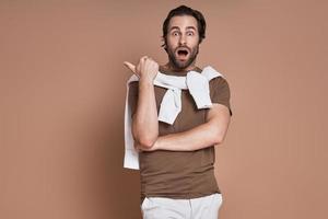 Shocked young man pointing away while standing against brown background photo