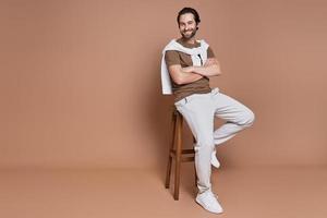 Fashionable young man keeping arms crossed and smiling while sitting against brown background photo