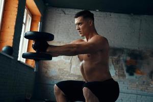 Concentrated young man standing in squatting position while training with dumbbell in gym photo