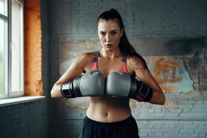 mujer joven confiada en guantes de boxeo mirando a la cámara mientras está de pie en el gimnasio