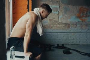 Young muscular man carrying towel on shoulders while sitting on the window sill in gym photo