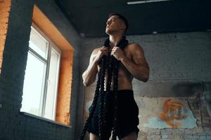 Handsome young man carrying battle rope on shoulders while standing in gym photo