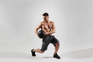 joven concentrado con cuerpo perfecto haciendo ejercicio con balón medicinal contra fondo blanco foto