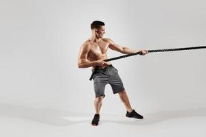 Concentrated young man with perfect body pulling a rope against white background photo